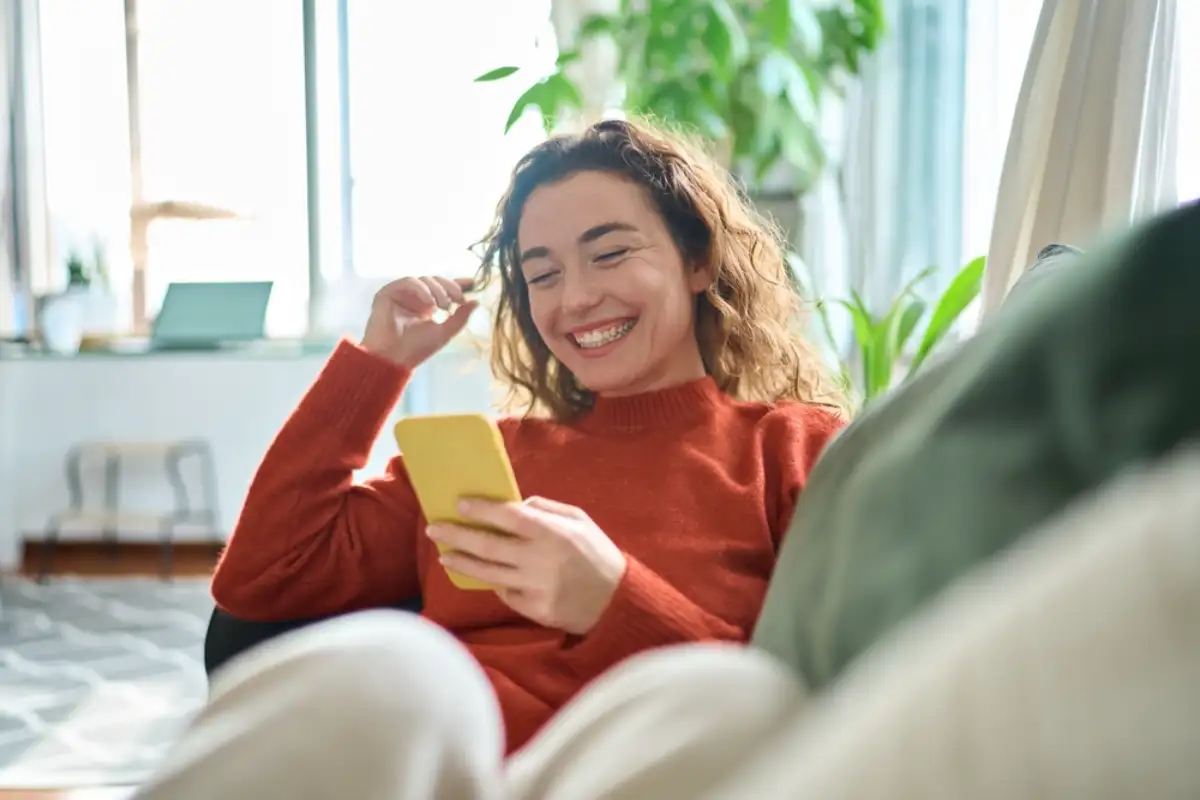 Happy relaxed young woman sitting on couch using cell phone