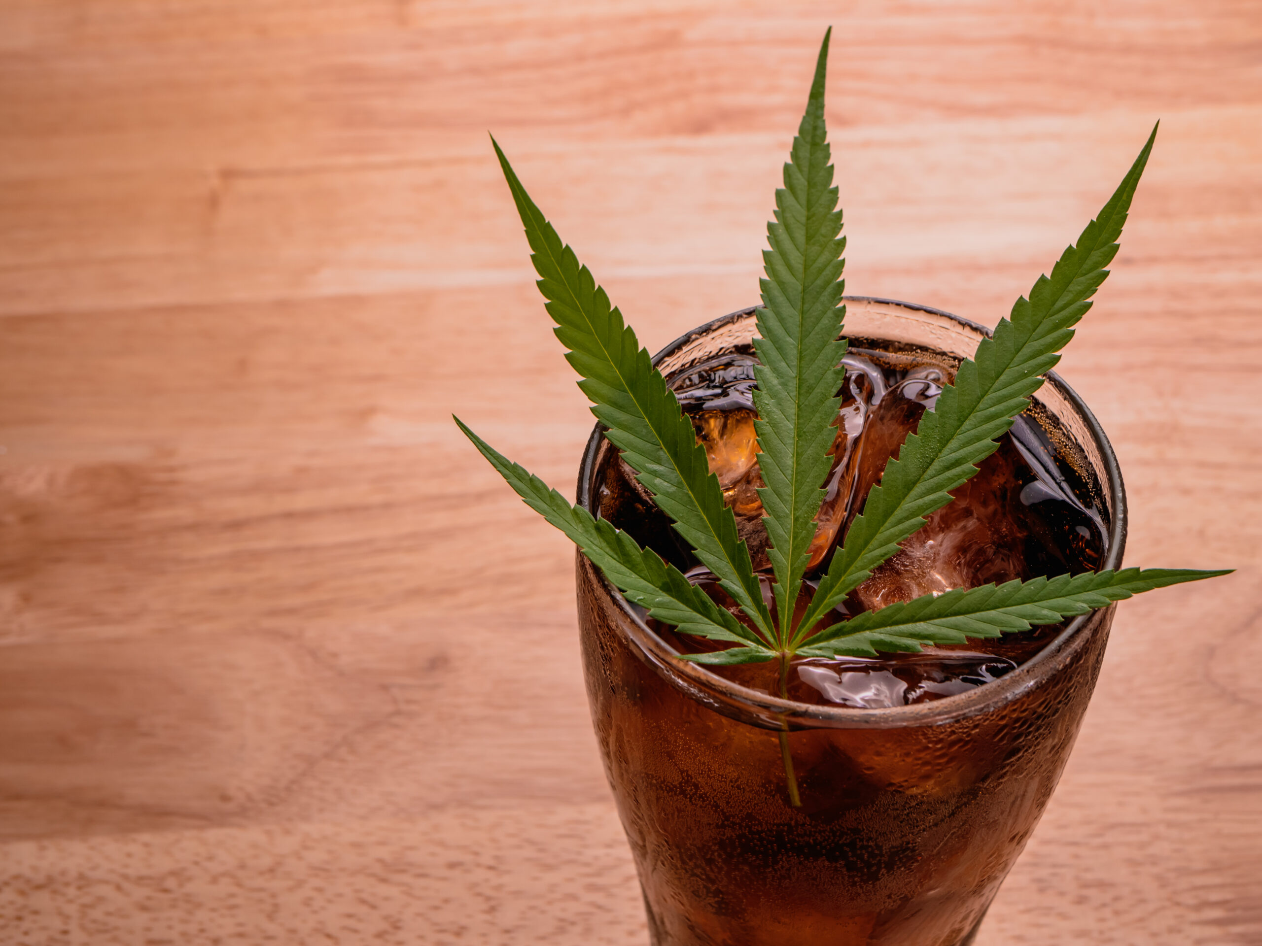 Glass of cola with ice cubes and cannabis leaf on the wooden tab